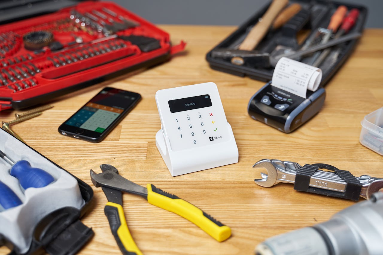 creative-02 Red and black toolboxes next to smartphone and modern cash registers composing with pliers and wrenches on beige wooden table in bright room