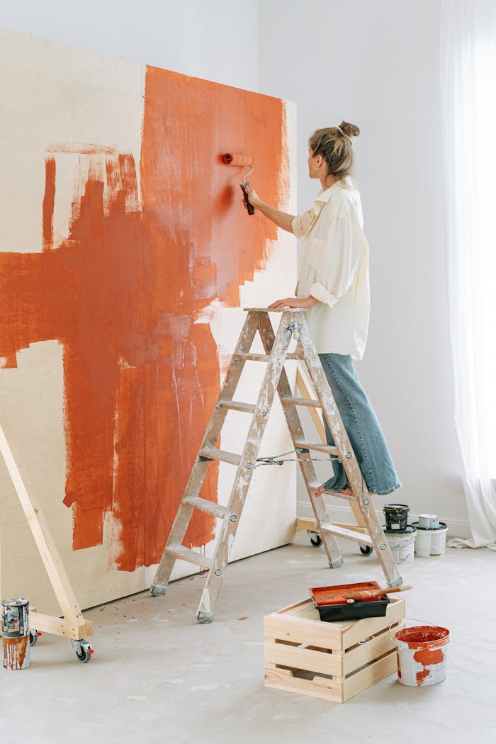 Services-01 A woman stands on a stepladder, painting a room's wall during home renovation.